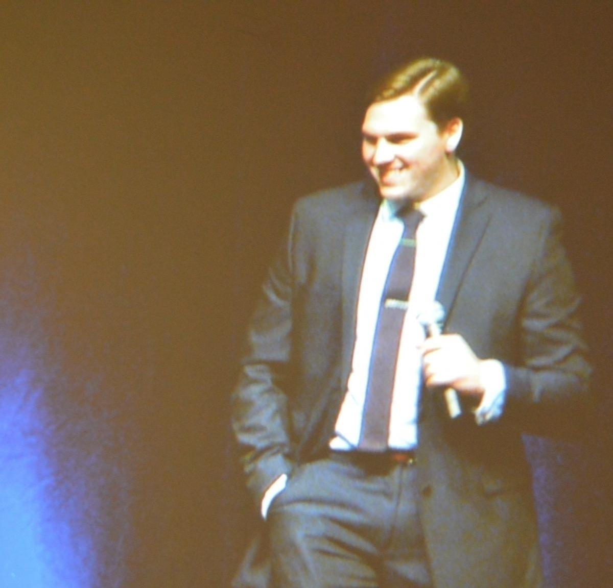 Man in suit standing on stage speaking into microphone.