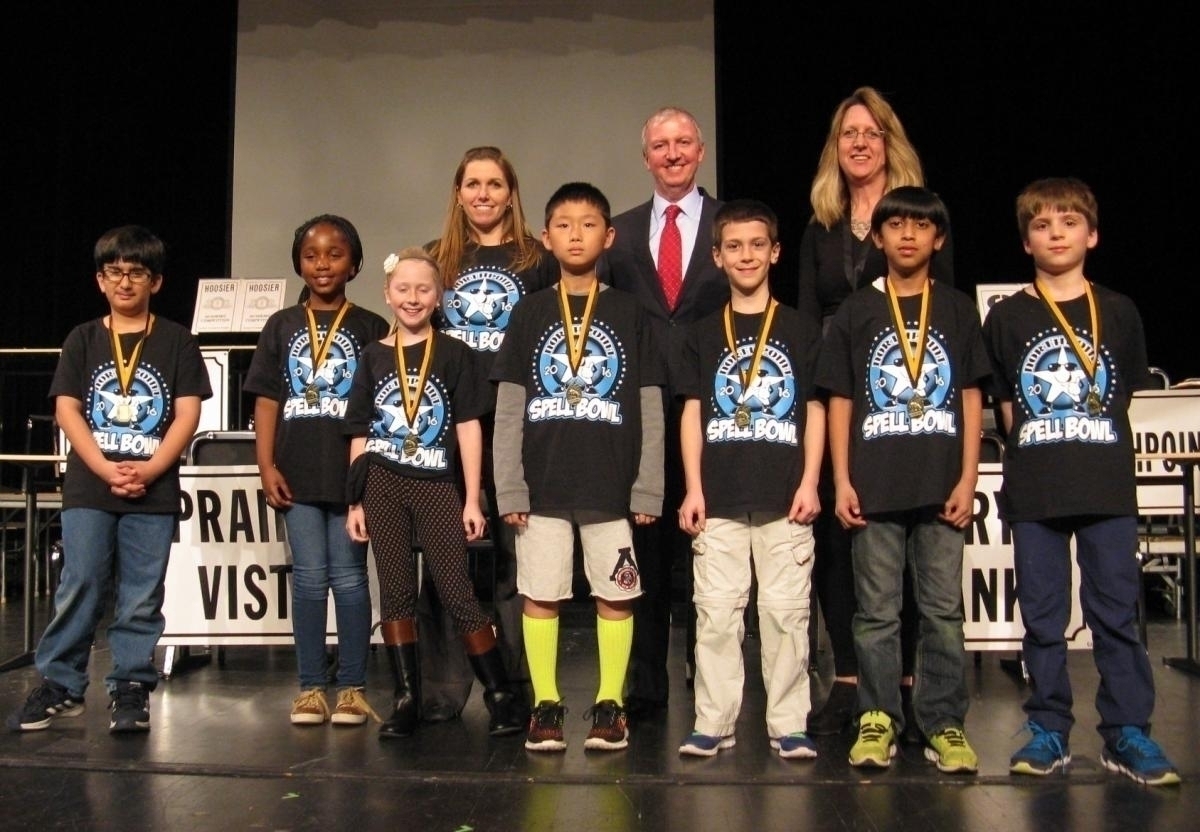 The group of children wearing black shirts with white lettering are standing on a stage in front of a podium.