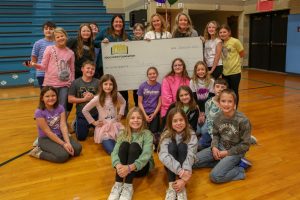 children in a gym surrounding a woman holding a giant check