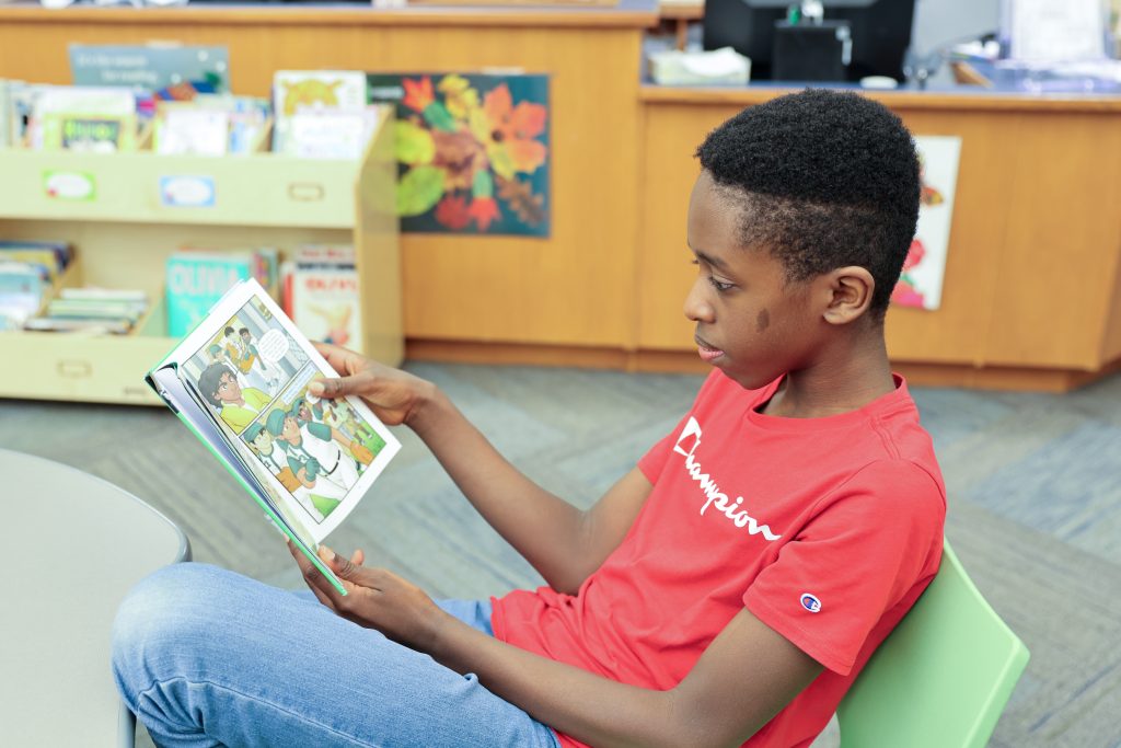 Boy in red shirt sitting on green chair reading a book.