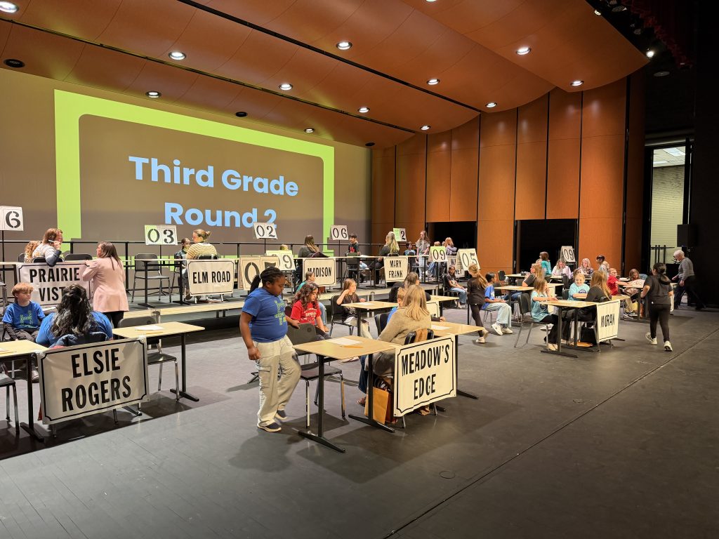 An indoor scene of a voting booth with people standing around tables displaying signs for various candidates and ballot