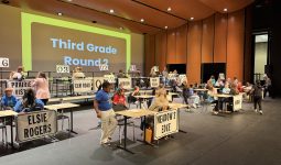 An indoor scene of a voting booth with people standing around tables displaying signs for various candidates and ballot