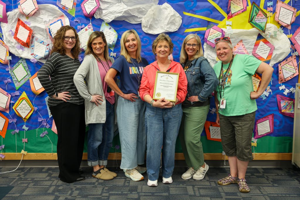 Five people posing in front of a bulletin board decorated with various items, including paper clouds and cut-out shapes,