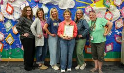 Five people posing in front of a bulletin board decorated with various items, including paper clouds and cut-out shapes,