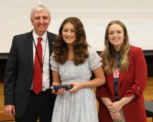 Three people standing on stage at an event, a young girl holding an award, smiling and posing with two adults, one man in