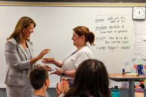 Two women in a classroom setting, one standing and handing something to another who is seated at a desk.