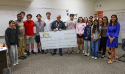 A group of people posing in a classroom setting, some holding a check, celebrating an achievement.