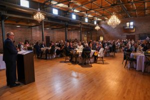 This is a photograph of an indoor event taking place in a large room with high ceilings and exposed brick walls. A man