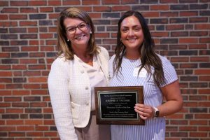 Two women posing together in front of brick wall; one holding plaque, smiling at camera.