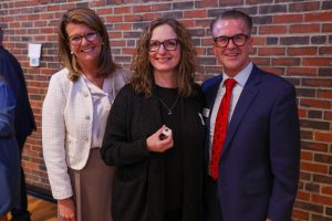 Three people posing for a picture at an event. A woman in a black shirt stands between two men; one is wearing glasses and a