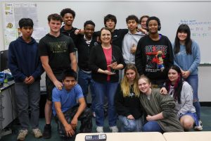 A teacher holding an award standing with a group of students in a classroom.