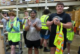 Group of people in a workshop setting, some clapping, others standing around, all wearing safety vests and hard hats, posing