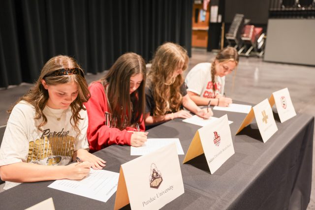 Five girls signing papers at a table in front of a podium.