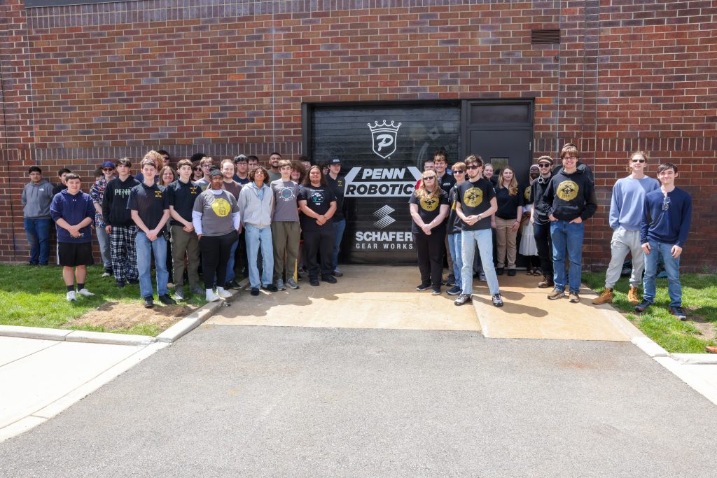 A group of people posing in front of a building with a sign that says "PENN ROBOTICS" and a logo for a robotics team.