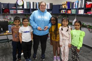 A teacher with a group of her young students standing in a room with books behind them.