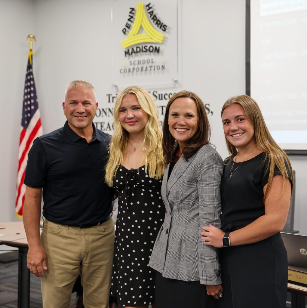 Four people posing together in front of a presentation screen, with one person standing behind the others and all smiling at