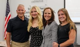 Four people posing together in front of a presentation screen, with one person standing behind the others and all smiling at