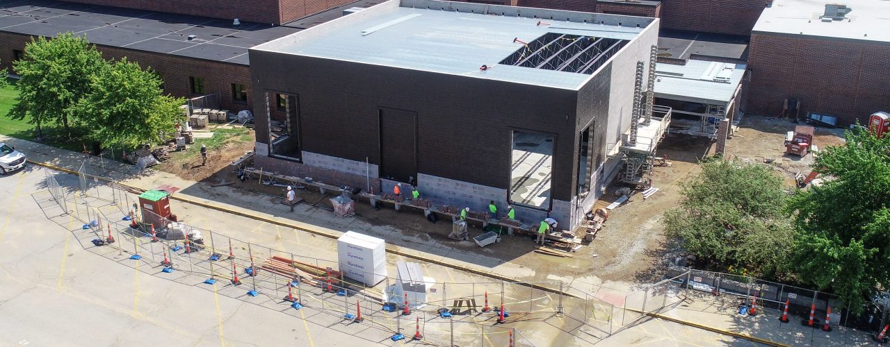Construction site in progress, featuring a large building with visible steel beams and scaffolding, surrounded by fencing