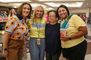 Four women posing together at an event, smiling and looking towards the camera. They are standing in front of a table with