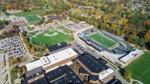 An aerial view of a school campus during autumn, showcasing the football field, parking lot, and nearby buildings.