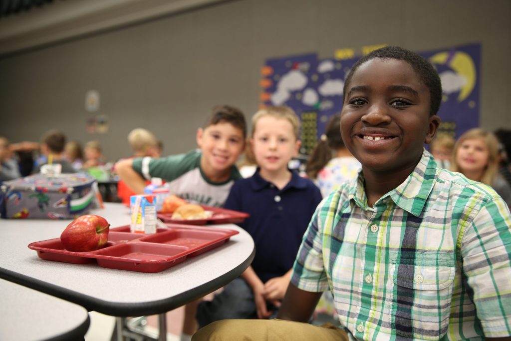 Young boy sitting at a table in a classroom, smiling towards the camera.