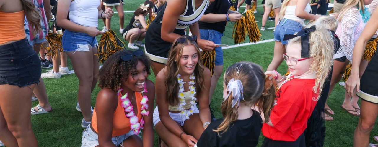 A group of cheerleaders, high school students, and younger students in a stadium setting, with some standing and others sitting on the grass talking and trading pom-poms.