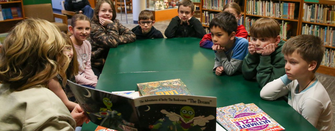 Children gathered around a table in a library setting, listening to an adult reading a book aloud.