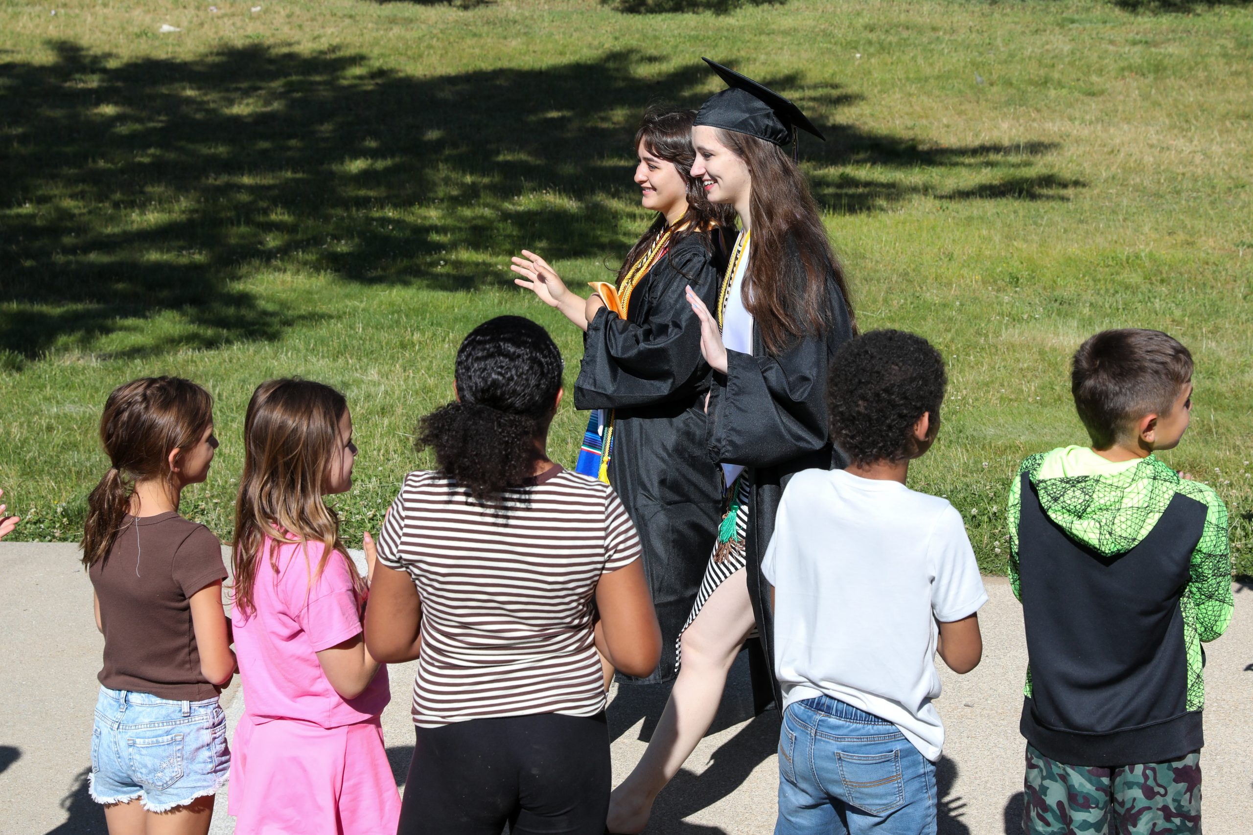 High school students in cap and gown walking near and waving to younger students