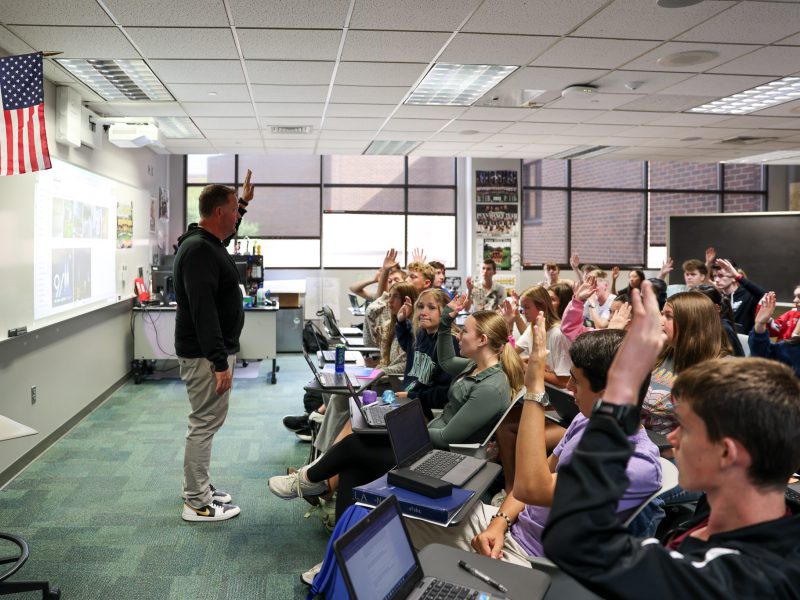 An educator in a classroom setting, presenting to a group of students who are seated in chairs and using laptops.