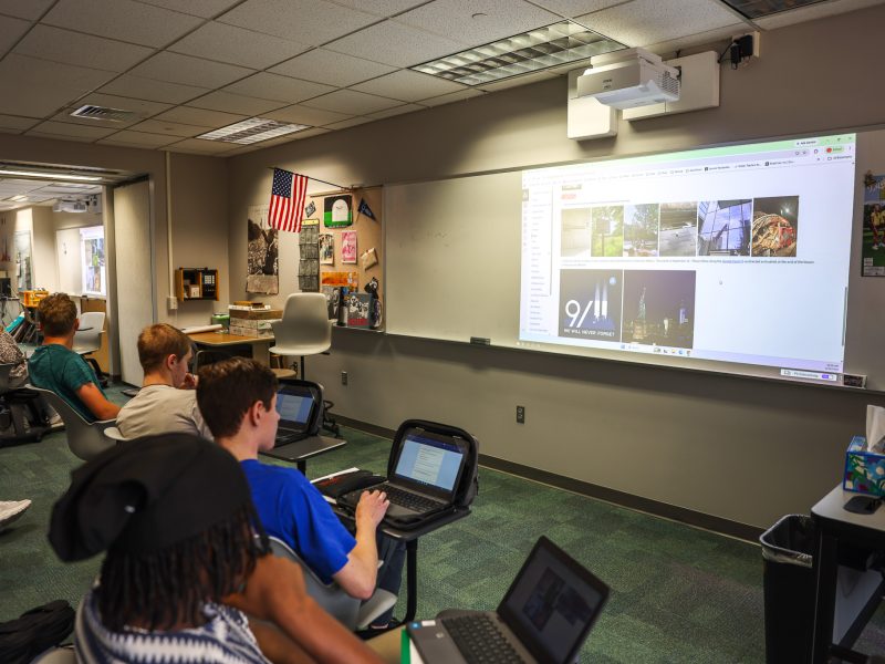 An educator in a classroom setting, presenting to a group of students who are seated in chairs and using laptops.