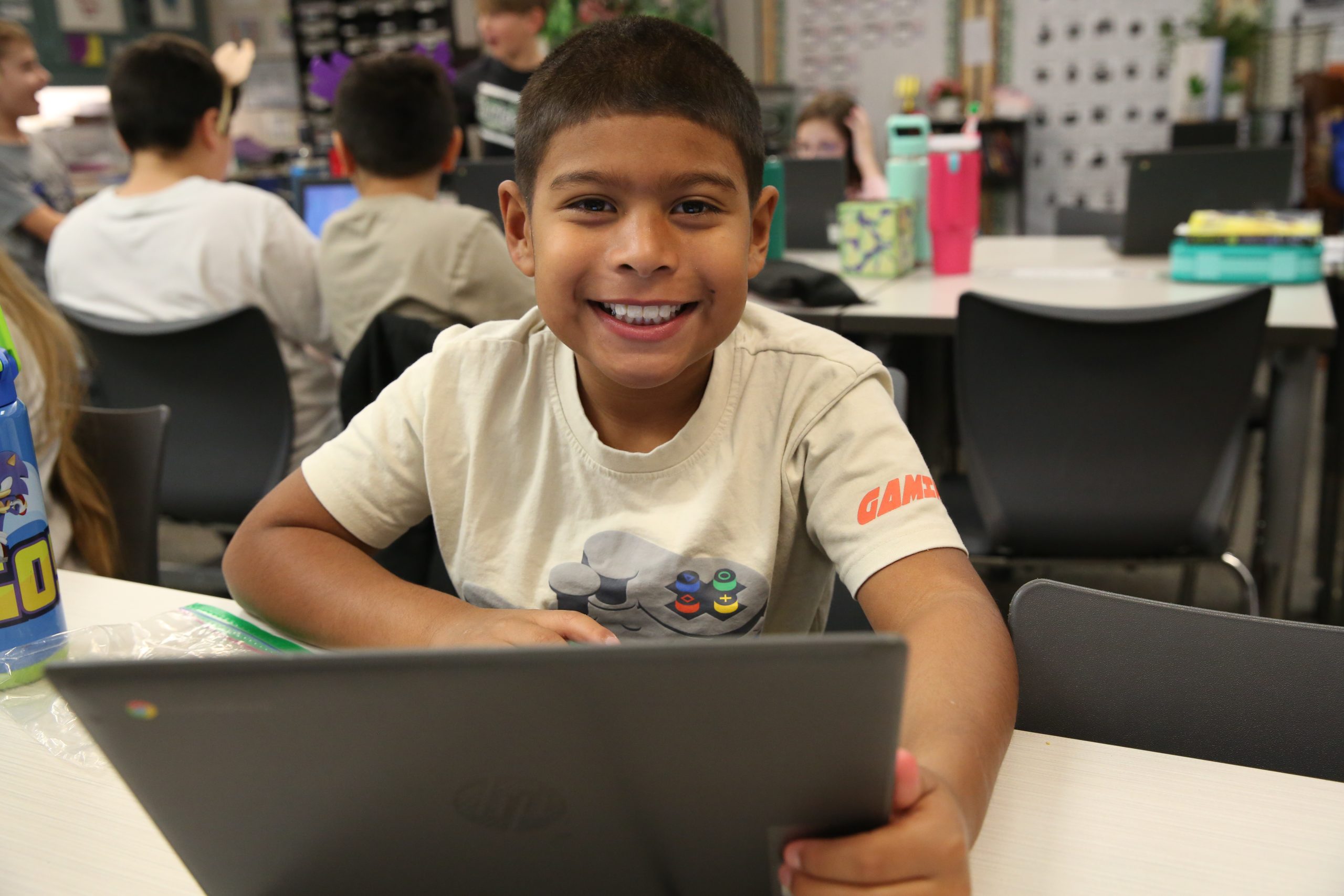 A young boy sitting at a table in a classroom, smiling at the camera, with a Chromebook in front of him.