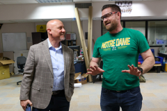 Two men in a room, one wearing glasses and a green shirt, the other in a suit. They are standing next to each other, engaged